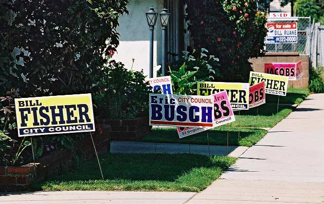 Photo showing three sets of campaign signs for Eric Busch (white), Sandra Jacobs (pink to attract the female voters), and Bill Fisher (yellow), all crowded on the same property.